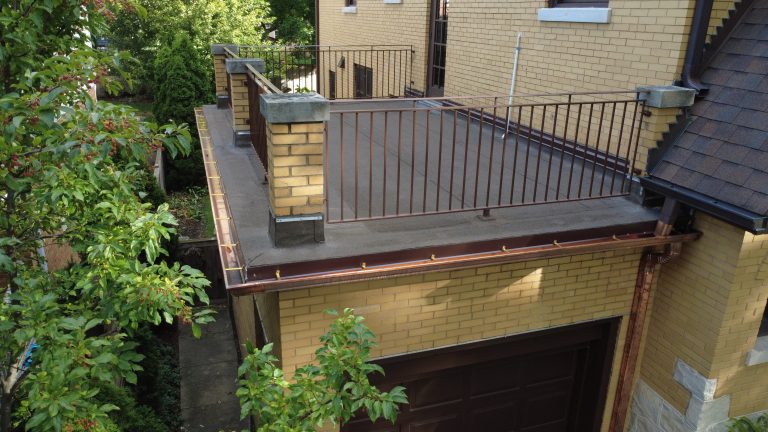 Flat roof deck over garage with new metal railings, fresh roofing surface, and copper gutters on a yellow brick home.
