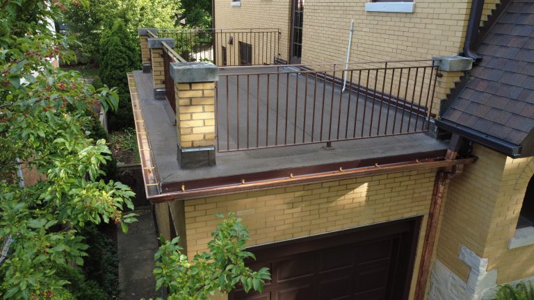 Flat roof deck above garage with metal railings, new roofing surface, and copper gutters on a yellow brick home.