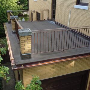 Flat roof deck above garage with updated roofing membrane, metal railings, and copper gutters on a yellow brick home.