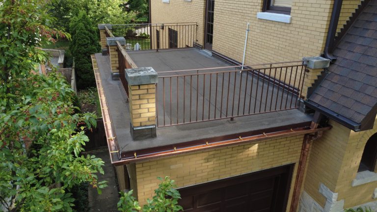 Flat roof deck above garage with updated roofing membrane, metal railings, and copper gutters on a yellow brick home.