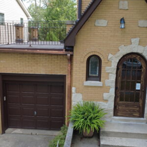 Front view of a brick home with an arched wooden door, updated flat roof deck above the garage, metal railings, and new copper gutters.