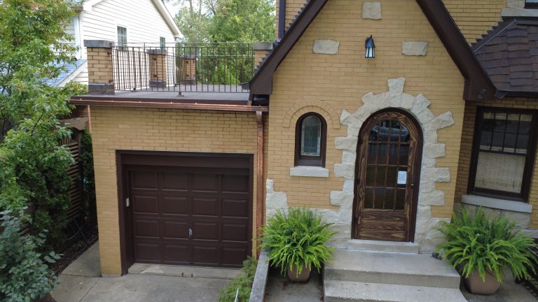 Front view of a brick home with an arched wooden door, updated flat roof deck above the garage, metal railings, and new copper gutters.