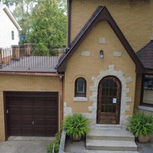 Front view of brick home with arched wooden door, restored flat roof deck above garage, metal railings, and new copper gutters.