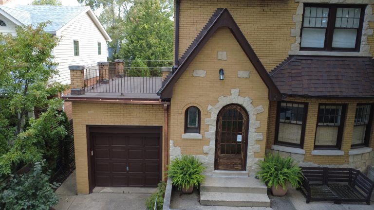 Front view of brick home with arched wooden door, restored flat roof deck above garage, metal railings, and new copper gutters.