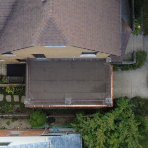 Top-down aerial view of a brick home with a restored flat roof deck, metal railings, copper gutters, and surrounding landscaping.