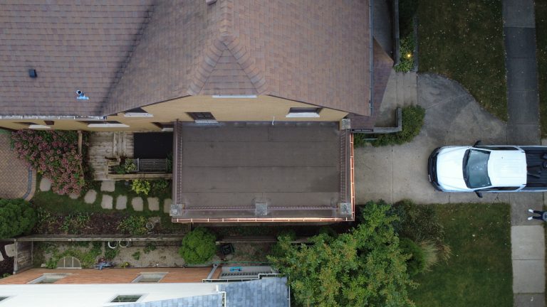 Top-down aerial view of a brick home with a restored flat roof deck, metal railings, copper gutters, and surrounding landscaping.