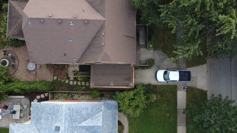 Aerial overhead view of a brick home with a restored flat roof deck, surrounding landscaping, patio area, and driveway.