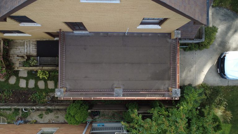 Aerial top-down view of a restored flat roof deck on a brick home with clean roofing membrane, metal railings, and copper gutters.