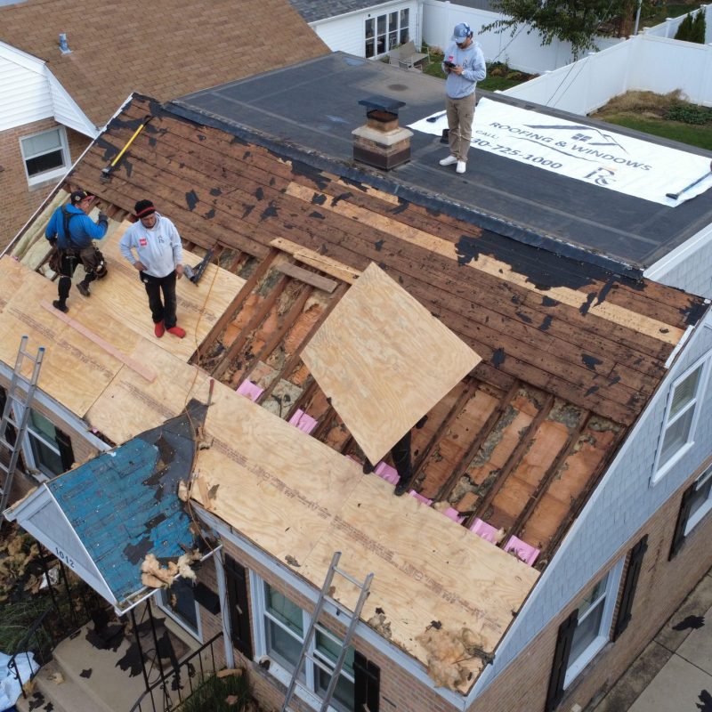Aerial drone view of a crew performing residential roof tear-off and replacement, exposing the decking and sheathing for repair before installing new roofing materials.