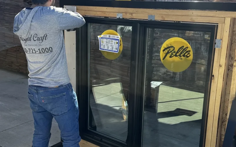 A worker from Royal Craft installs a black-framed Pella window outdoors on a sunny day.