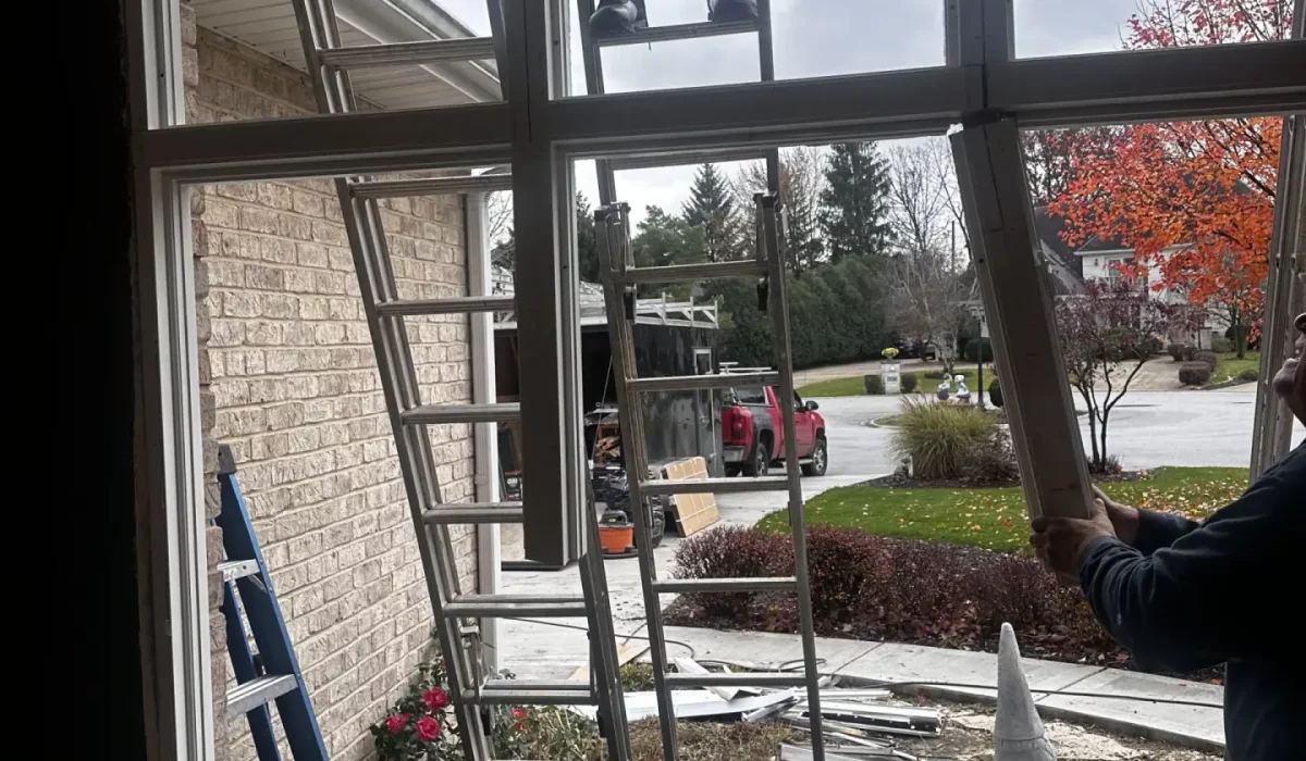 Technician removes old window frame during window replacement for Hinsdale home on overcast day with ladders setup.