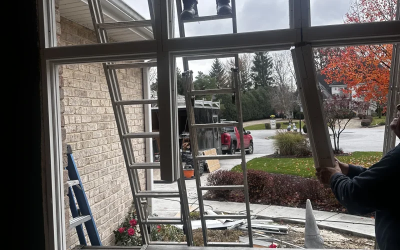 Technician removes old window frame during window replacement for Hinsdale home on overcast day with ladders setup.