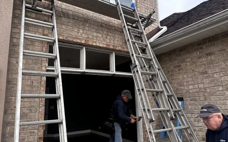 Two installers work on a large window replacement for a brick home, with ladders and scaffolding set up for access.