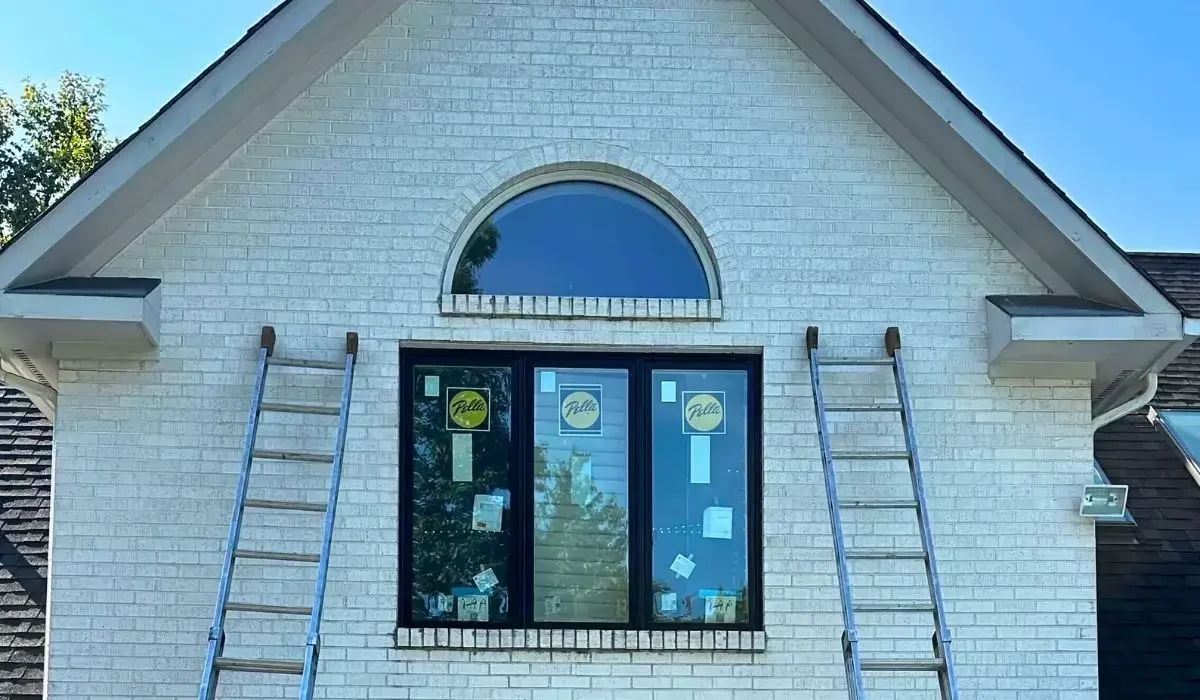 Second-story brick home with new window replacement in progress using ladders and energy-efficient black frame windows.