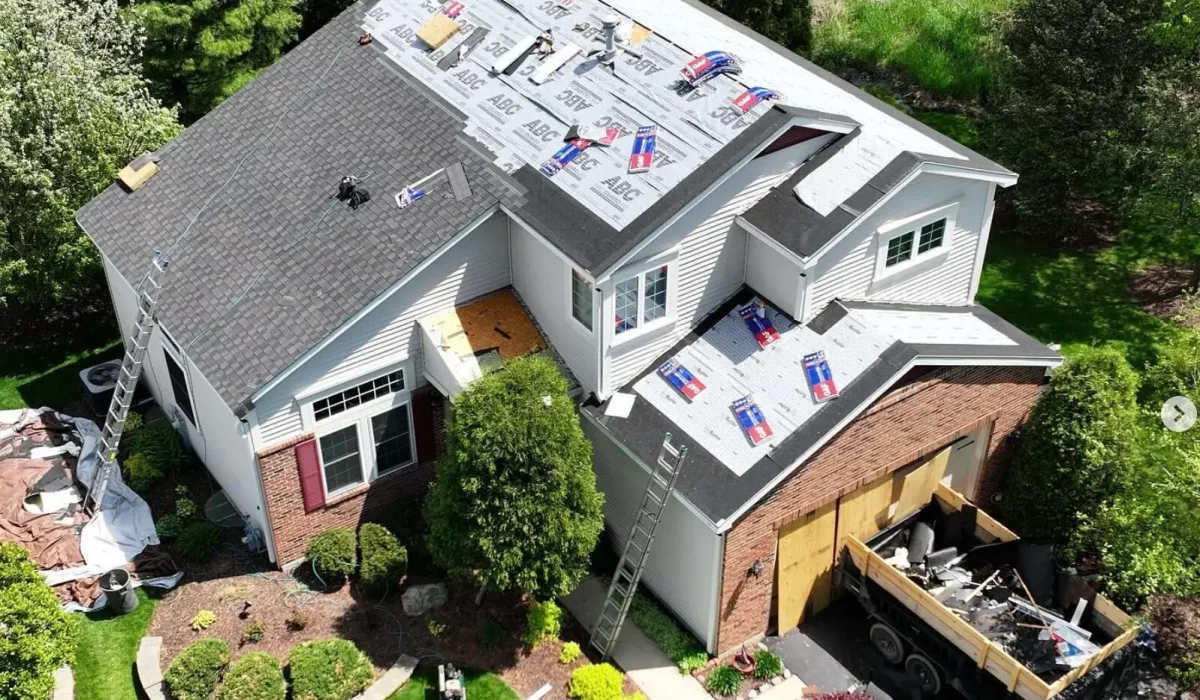 Roofing companies replace shingles on a two-story home, with equipment, ladders, and a debris truck nearby.