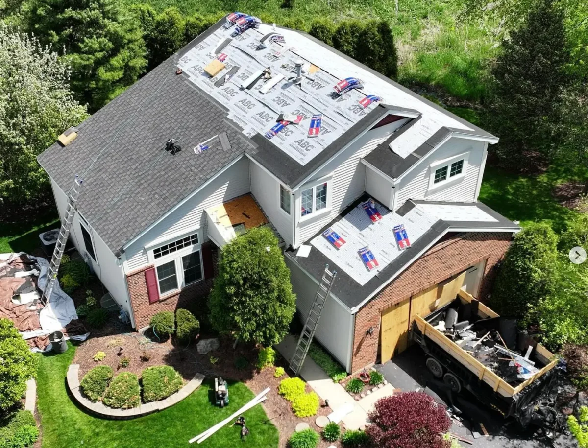 Roofing companies replace shingles on a two-story home, with equipment, ladders, and a debris truck nearby.