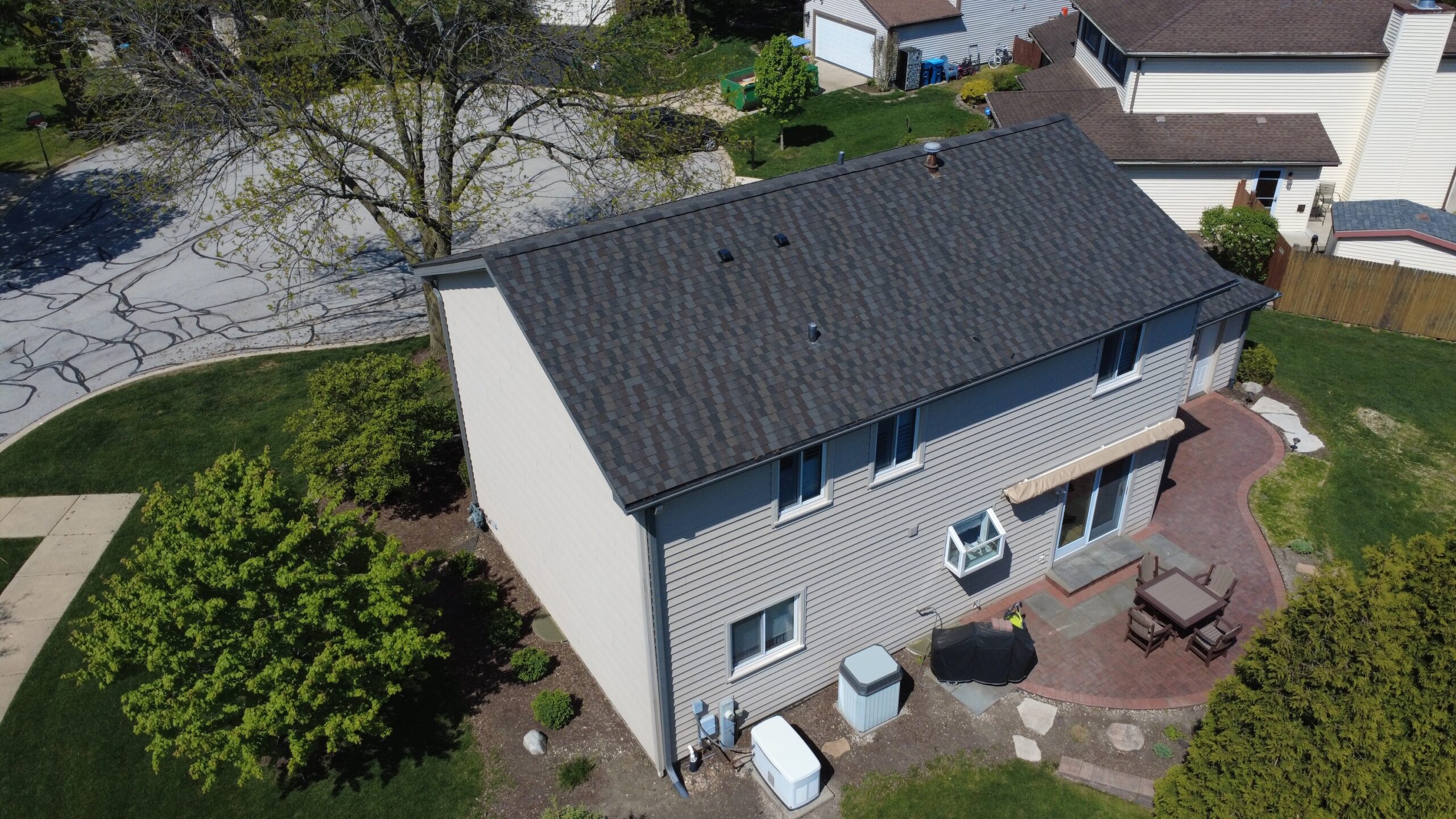 Aerial view of a two-story home with a newly installed dark gray asphalt shingle roof and brick patio.