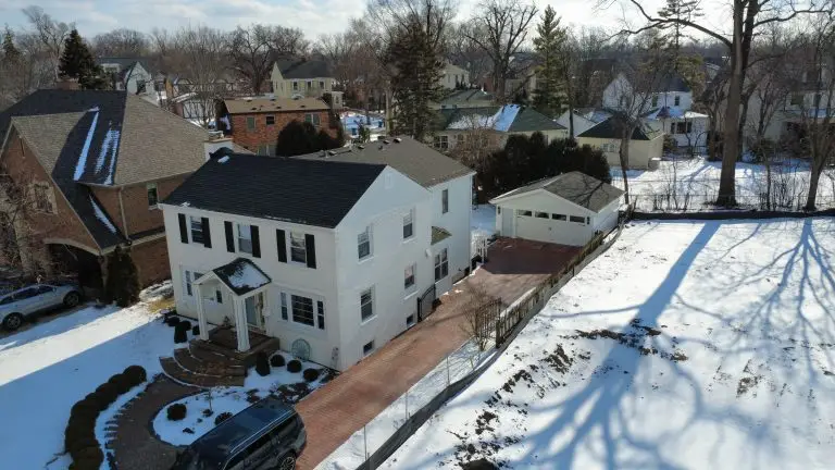 Winter drone view of a black shingle roof on a white colonial home with a brick paver driveway
